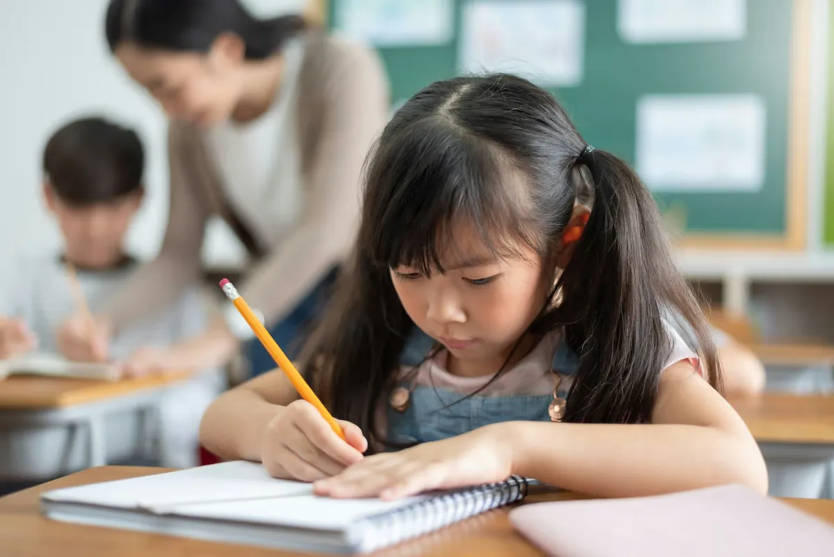 A young student concentrates on writing at her desk in a classroom
